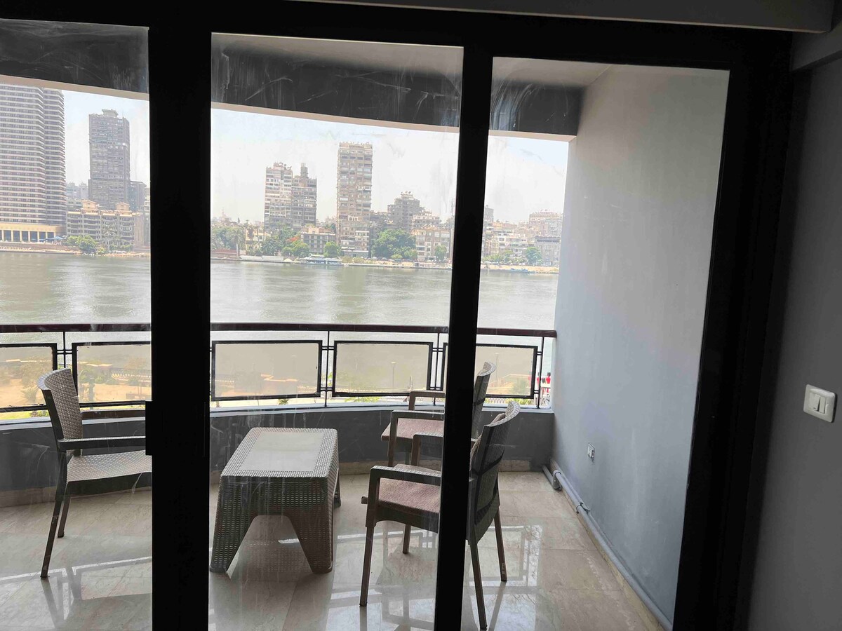 A balcony area features a glass door leading to a view of the Nile river. Two chairs and a small table are positioned on the polished floor, allowing for relaxation while enjoying the river scenery and the city skyline in the background.