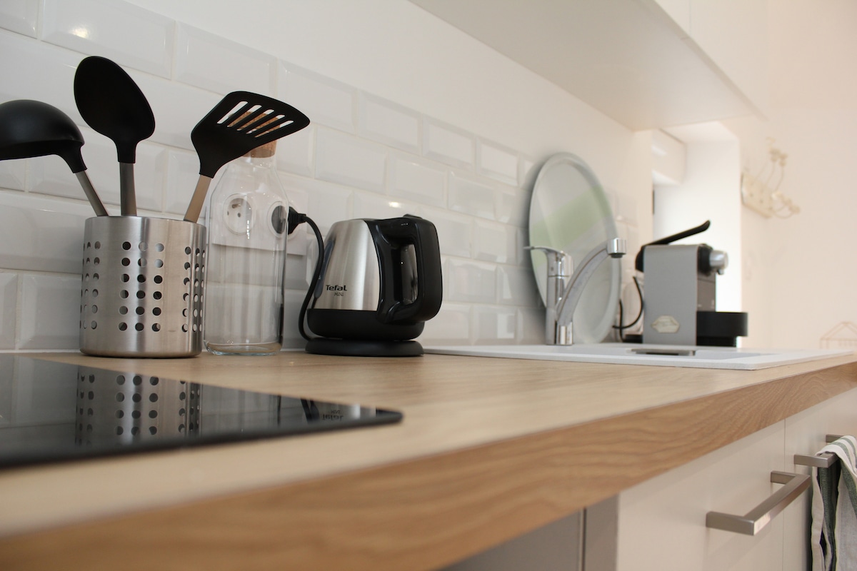 A compact kitchen workspace features a wooden countertop with cooking utensils, a glass water bottle, and a small electric kettle. An espresso machine and a mirror are positioned beside an induction cooktop, providing essential amenities for meal preparation.