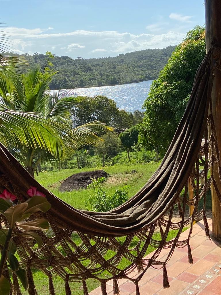 A hammock is shown gently swaying in the breeze, offering a view of lush greenery and shimmering water in the distance. Tall palm trees frame the scene, with sunlight illuminating the landscape and casting soft shadows on the ground.