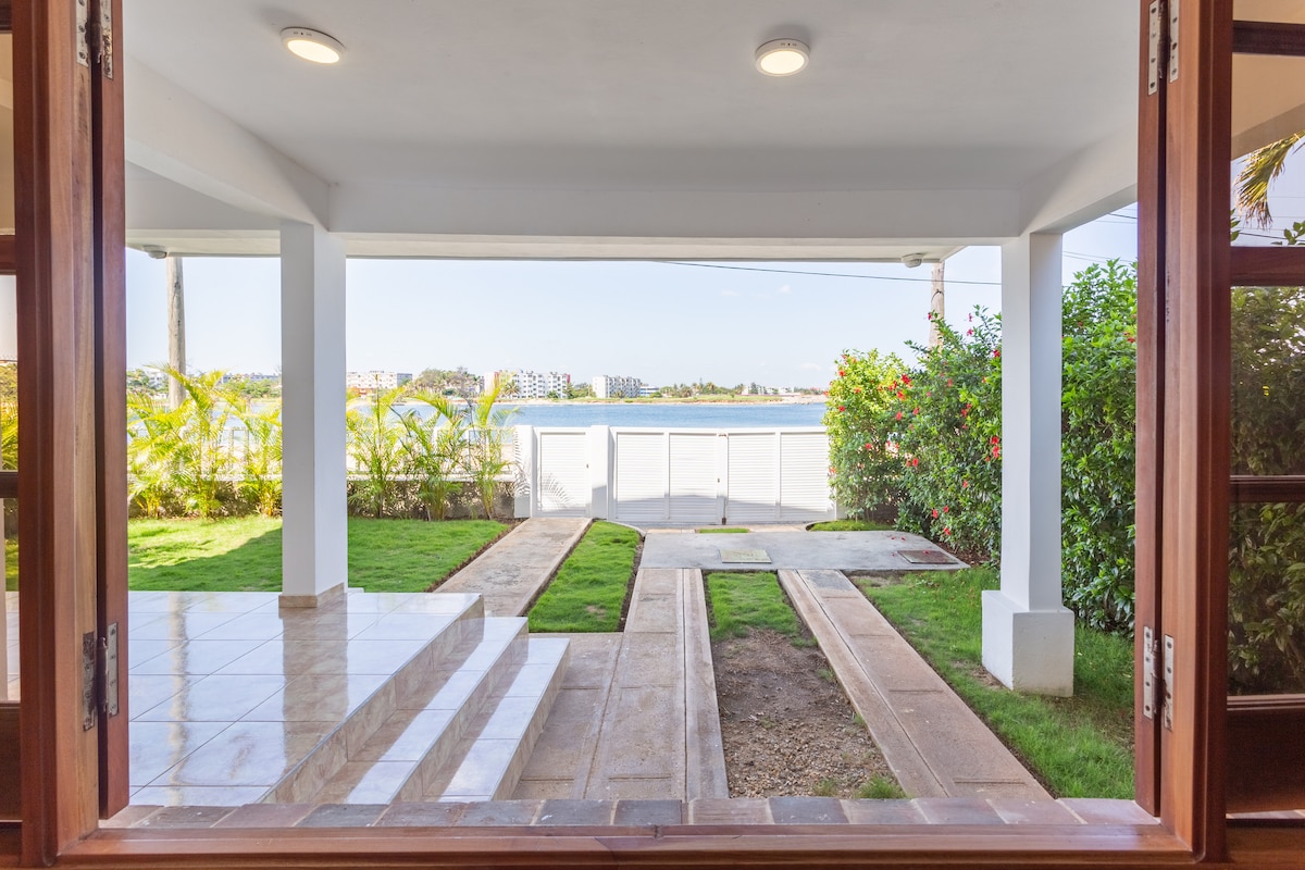 A view from the indoor space frames a green garden area leading to the waterfront. The pathway is bordered by neatly trimmed grass and vibrant foliage, with a glimpse of a serene waterway in the background. Large sliding doors invite natural light into the interior.