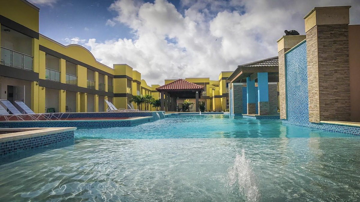 A communal pool area is seen, featuring a large, inviting blue water pool surrounded by lounge chairs. Bright yellow buildings line the perimeter, with a shaded area constructed of wood and stone visible in the background. Gentle splashes can be observed near the water's surface.