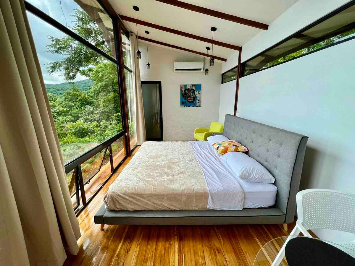 A spacious sleeping area features a large bed with soft linens and decorative pillows. Floor-to-ceiling windows provide views of the surrounding greenery, while warm wood flooring contributes to the serene ambiance. Modern light fixtures hang above, adding to the stylish decor.