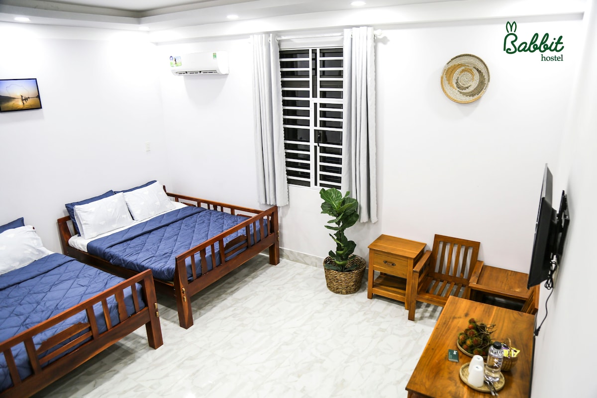 The room features two wooden beds with blue quilts and white linens. A small wooden table and chairs are positioned in one corner, while a potted plant adds a touch of greenery. Natural light enters through the window with white curtains.