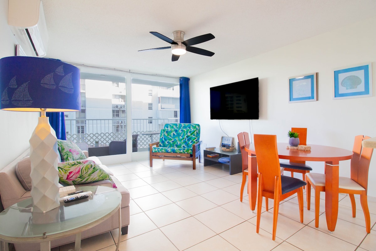 A bright living and dining area is featured, showcasing a ceiling fan and large windows allowing natural light. A comfortable seating arrangement includes a colorful sofa and armchair. A round dining table with orange chairs is positioned nearby, complementing the design of the space.