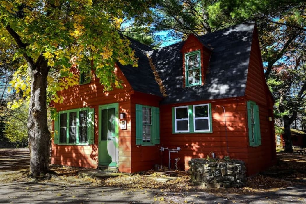 A charming rustic cabin is shown with a vibrant red exterior complemented by green shutters. Large windows invite natural light, and the surrounding trees display autumn foliage, enhancing the serene atmosphere. The stonework at the base adds a touch of craftsmanship.