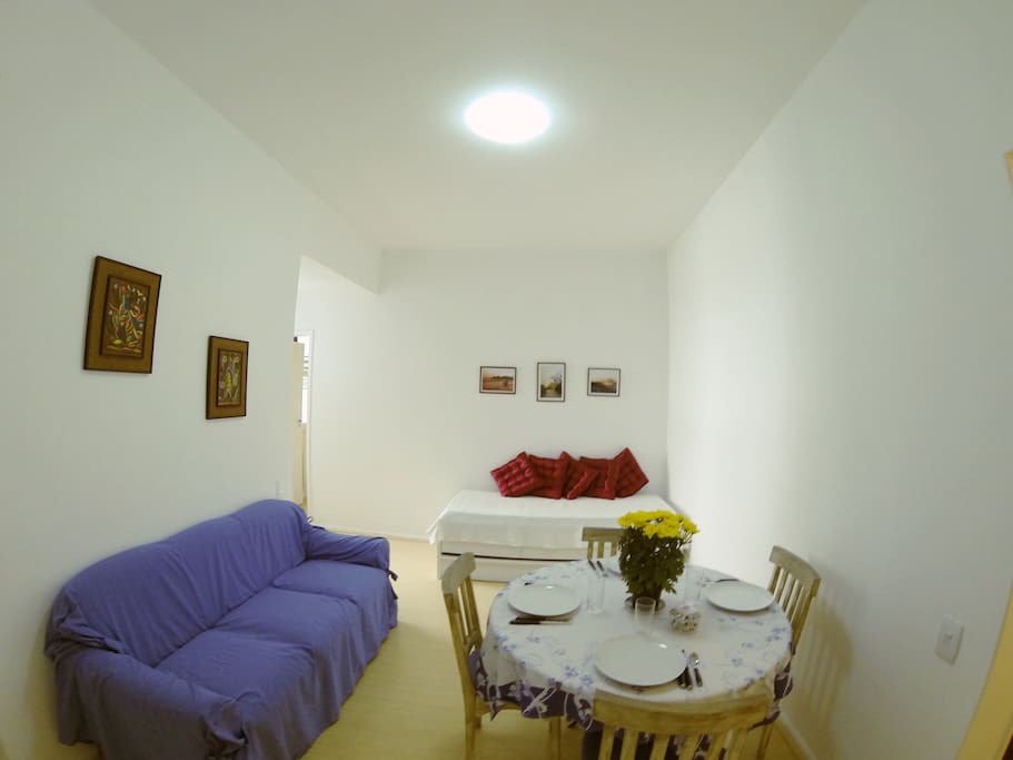Sala clara e arejada com mesa com quatro cadeiras, sofá de três lugares e bi-cama.