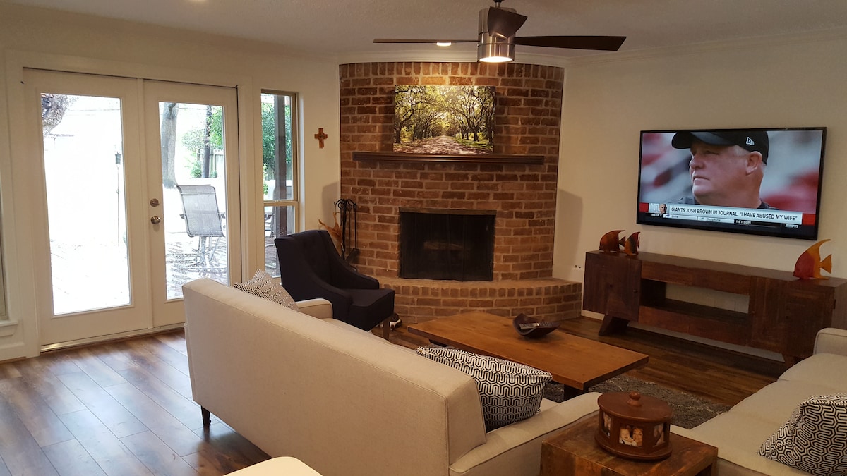 A living area is presented with a brick fireplace as a focal point, accompanied by a flat-screen TV. Seating includes a light-colored sofa and a dark armchair. Large glass doors lead to an outdoor space, enhancing the openness of the room with wood flooring throughout.