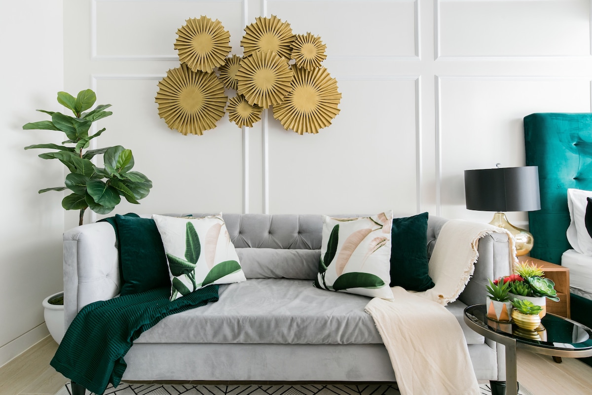 A plush gray sofa is complemented by decorative cushions featuring tropical leaf patterns. A muted throw blanket is draped casually over one side. Above the sofa, a set of golden wall art pieces adds a touch of elegance, while a potted plant contributes to the serene atmosphere.