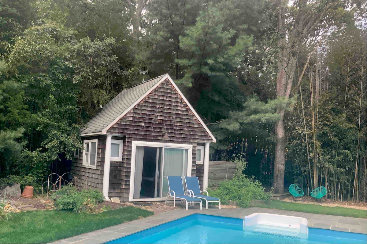 A charming guesthouse is situated near a tranquil pool. The exterior displays natural wood shingles, with large glass doors inviting light inside. Two blue lounge chairs are positioned by the pool, surrounded by lush greenery and a bamboo forest, creating a serene atmosphere.