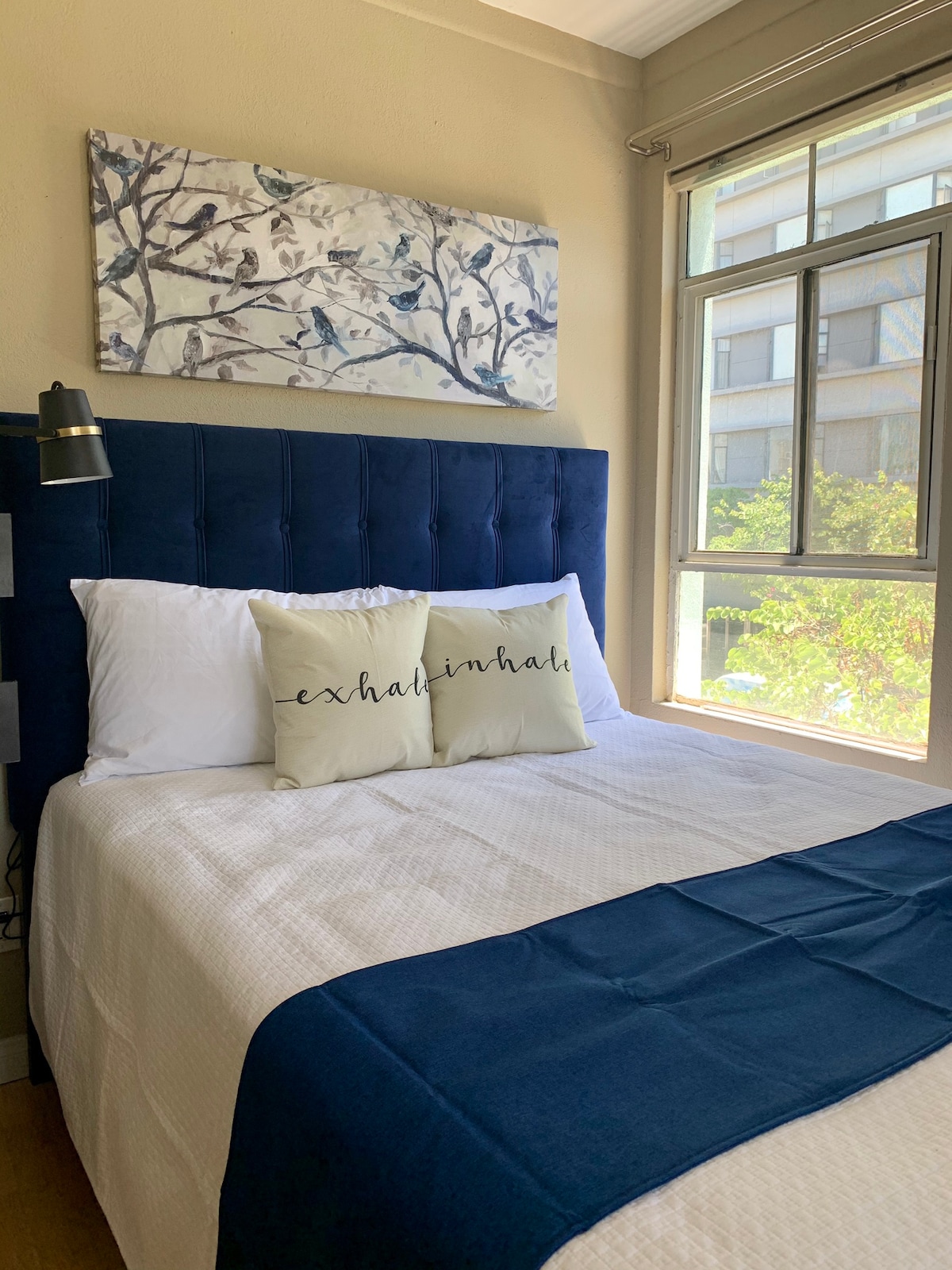 A cozy sleeping area features a plush bed with white linens and a navy blue throw. Two decorative pillows add a personal touch. Natural light is provided by the large window, framed by a light curtain and overlooking vibrant greenery.