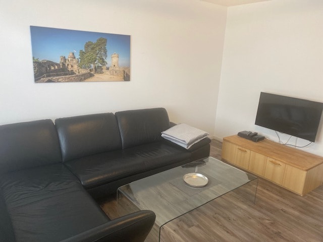 A spacious living area features a dark leather sofa, complemented by a glass coffee table atop a wooden floor. A television is situated on a wooden cabinet, and a framed landscape artwork adorns the wall, depicting a historic castle.