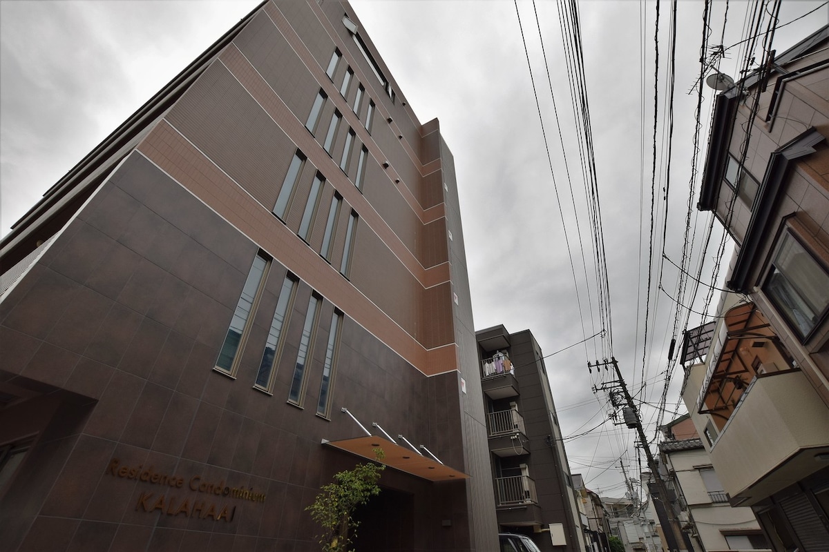 The exterior of a multi-story building is presented, showcasing a modern design with a dark facade and large glass windows. A small green plant is positioned near the entrance, while power lines and neighboring buildings are visible in the surrounding area. A cloudy sky fills the background.