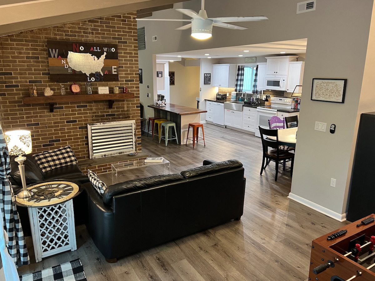 An inviting living area features a black leather sectional sofa, complemented by a rustic coffee table. A decorative wall with the U.S. map design adds character, while the open layout reveals a modern kitchen and dining space with colorful stools, enhancing the communal atmosphere.