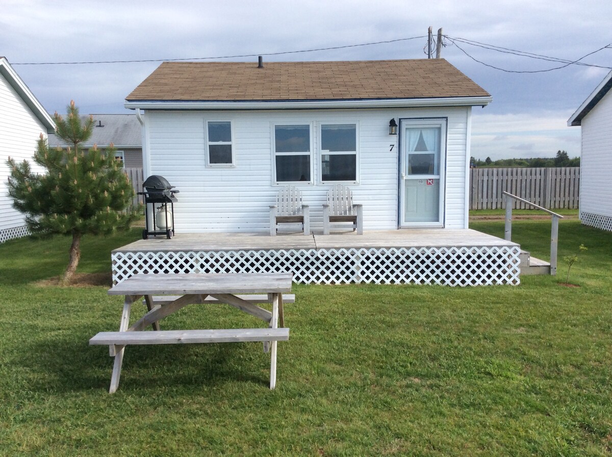 A waterfront cottage is showcased with a simple wood deck. Two chairs are positioned on the deck, overlooking the grassy area. A picnic table is visible in front of the cottage. A barbecue grill is located to the side, under a clear sky.