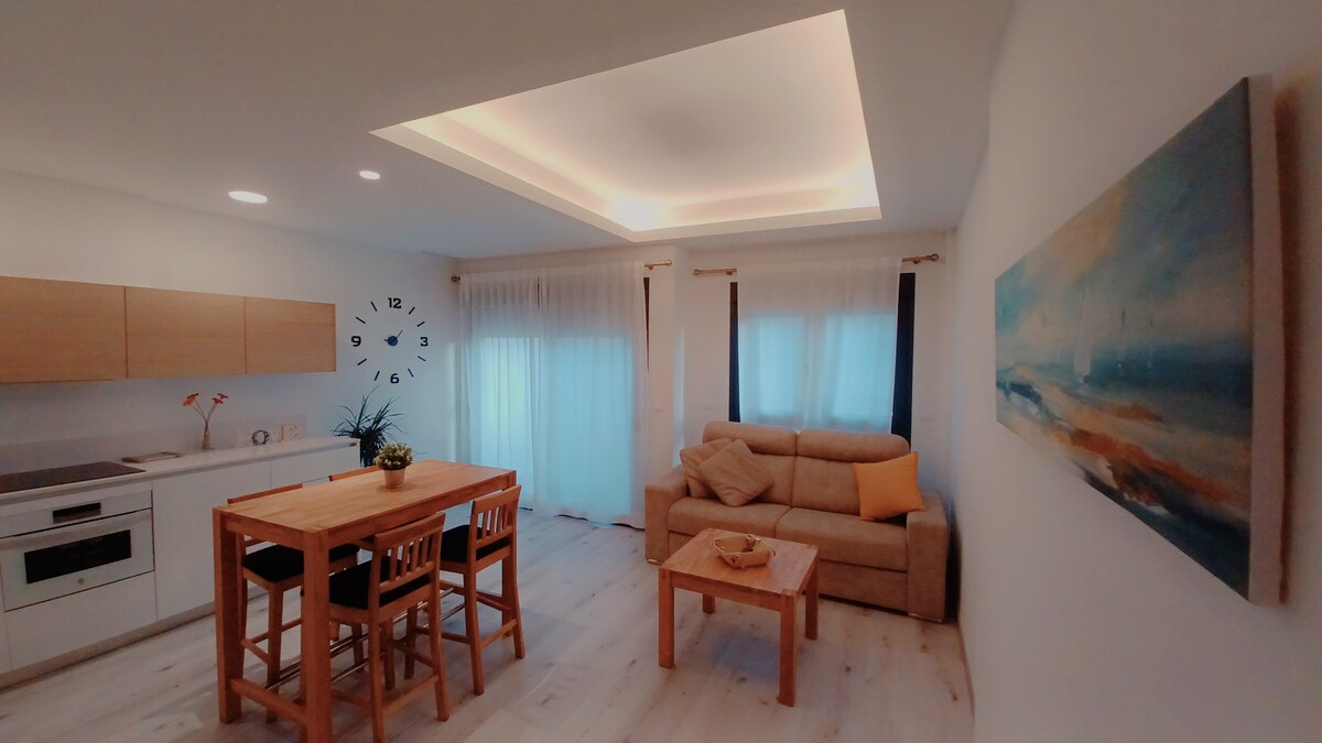 The open living area features a modern kitchen with light wood cabinetry and a dining table surrounded by four chairs. A comfortable sofa is positioned nearby, complemented by a small side table. Soft, diffused lighting creates a warm ambiance, and large windows allow ample natural light.