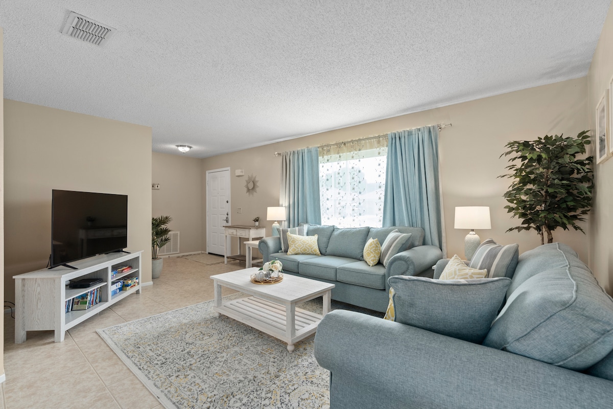 A comfortable living area is highlighted by soft blue furnishings and a light-colored area rug. A smart TV is positioned on a white media console, while natural light filters through sheer curtains. Potted plants and lamps add a touch of warmth to the space.