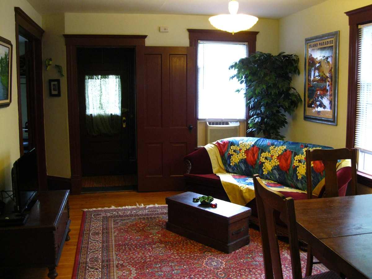 A welcoming living area is presented with a vibrant flower-patterned quilt draped over a comfortable couch. A wooden coffee table sits in front, and natural light filters through the front window, illuminating the space. Wooden accents and greenery enhance the inviting atmosphere.