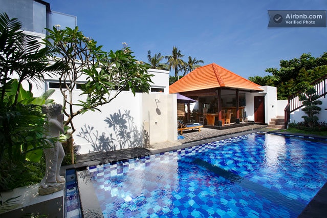 A vibrant swimming pool is surrounded by lush greenery and modern architecture. A traditional pavilion with a red roof and dining area is visible in the background, creating a harmonious blend of indoor and outdoor spaces. Sunlight reflects off the pool's surface.