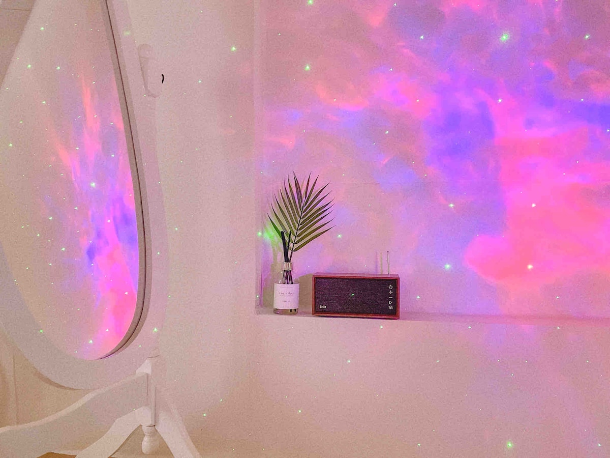 A modern space features a uniquely shaped mirror reflecting a colorful light display on the wall. A small plant is placed on the window sill, alongside a minimalist speaker. The atmosphere is filled with soft, ambient lighting creating a calming vibe.