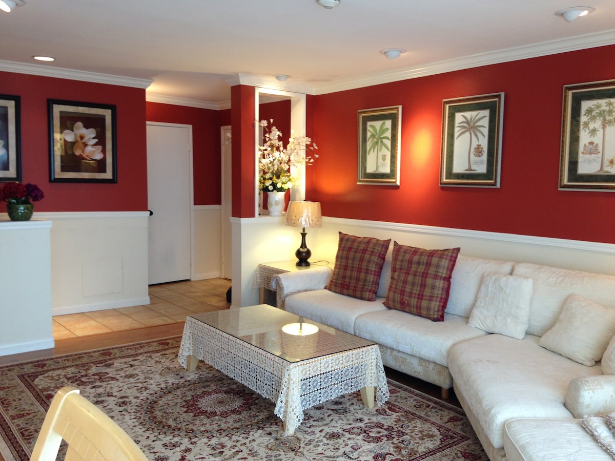 A living area is showcased with a white sectional sofa adorned with plaid and textured cushions. A patterned area rug covers the tiled floor, and a round table with a decorative tablecloth sits in front. Artwork is displayed on the red walls, accompanied by a lamp and floral arrangements.