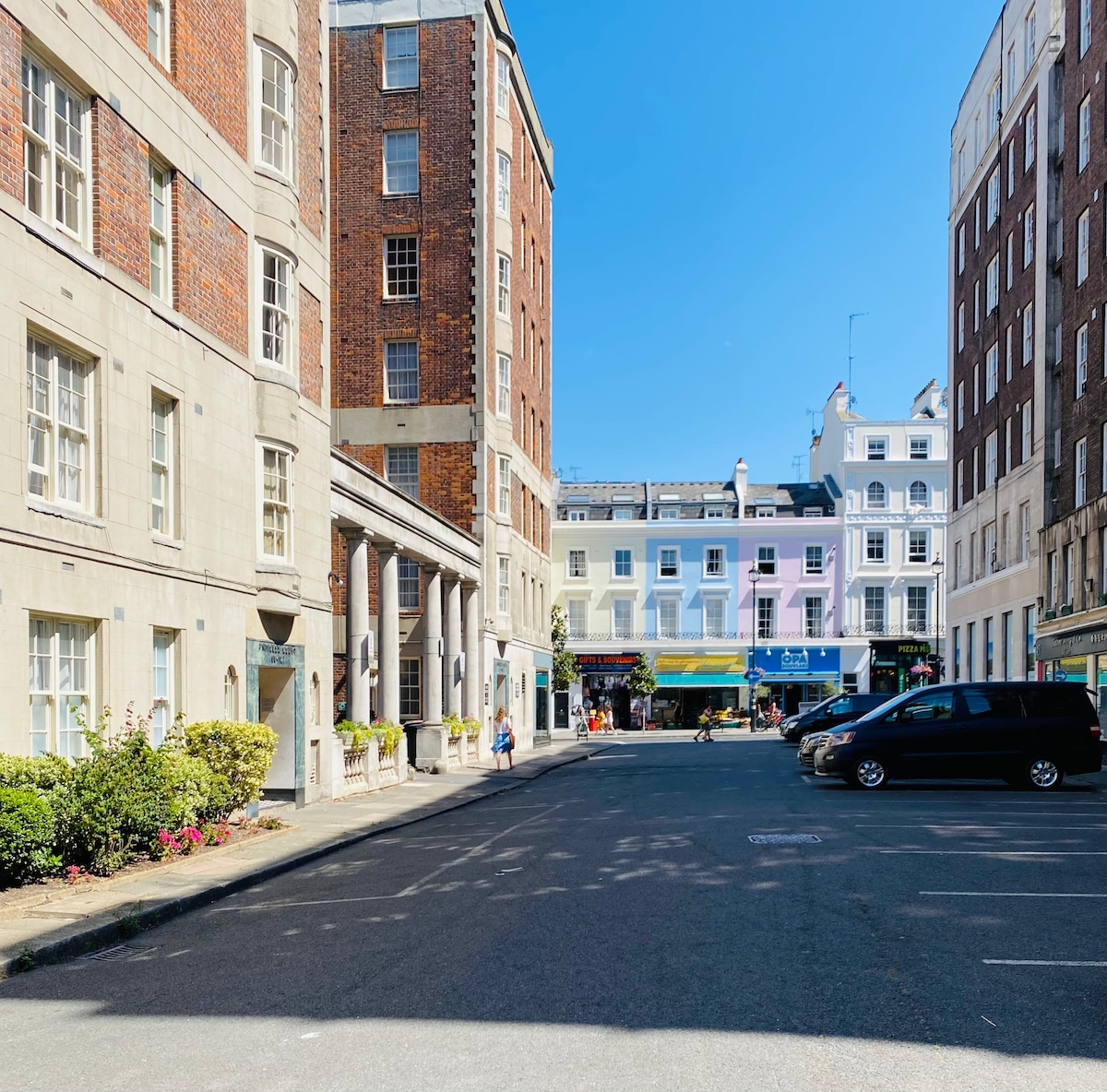 A view of a sunny street framed by multi-story buildings, featuring a mix of brick and smooth façades. Potted plants and flower beds are positioned along the walkways, while parked cars line the street. People are seen walking, suggesting a vibrant neighborhood.