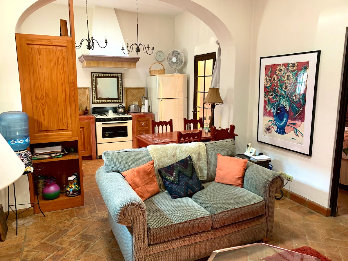 A cozy living area features a comfortable green sofa with decorative pillows, complemented by a wooden dining table and chairs. The well-equipped kitchen, visible in the background, includes a stove, refrigerator, and various appliances, with natural light entering through nearby windows.