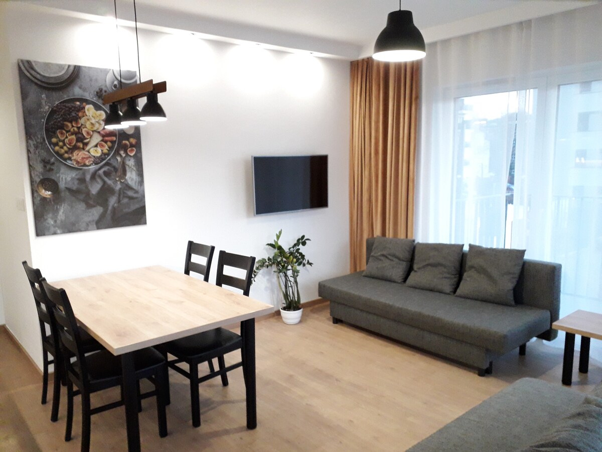 A modern living area features a wooden dining table with six chairs and a gray sofa. A wall-mounted television is positioned opposite the sofa. Natural light enters through large windows, complemented by soft curtains. A decorative plant is placed in the corner, adding a touch of greenery.