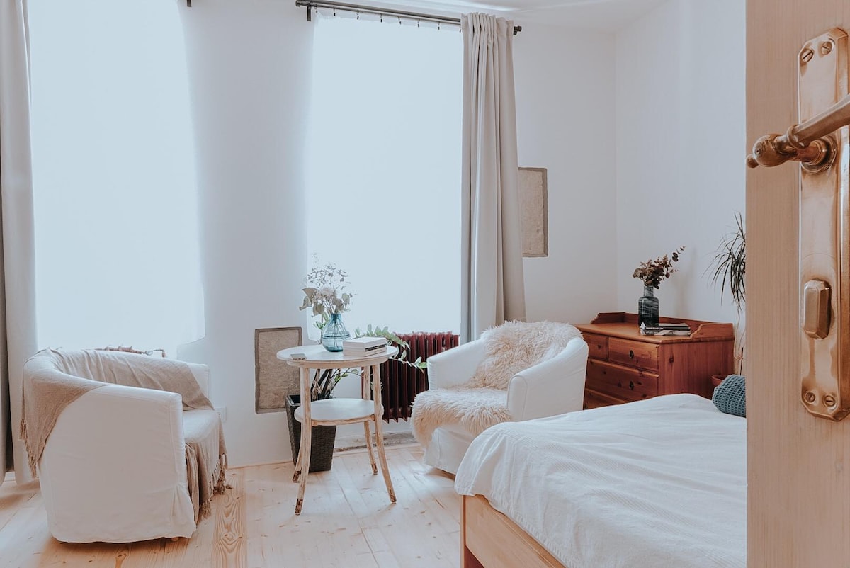 A bright and airy room is featured, showcasing two white armchairs and a small round table. Natural light enters through large windows, highlighting the wooden flooring. A simple dresser is visible, accompanied by decorative plants and vases adding a touch of greenery.