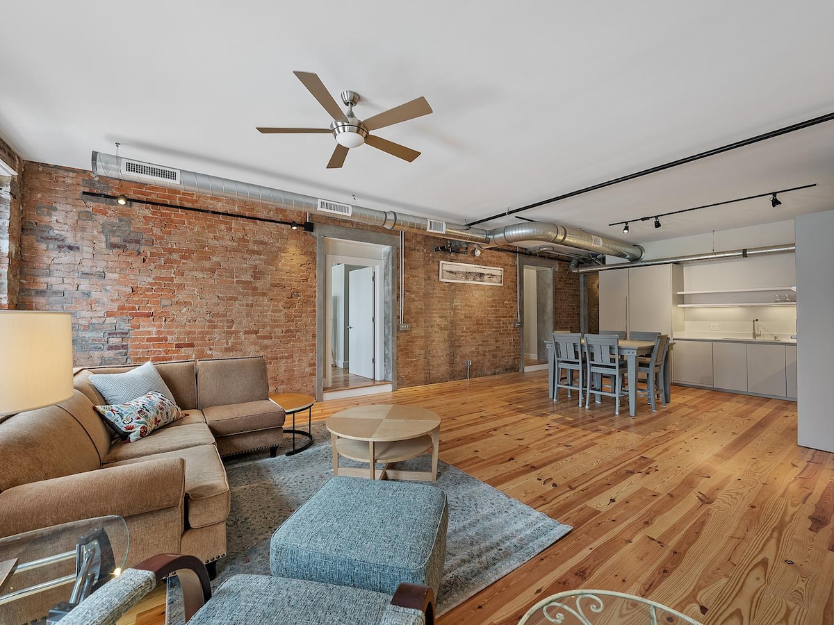 An open living space features warm hardwood flooring and exposed brick walls. A comfortable sofa is complemented by a coffee table and a cozy area rug. A dining table is arranged nearby, and a modern kitchen is visible in the background, enhancing the spacious atmosphere.