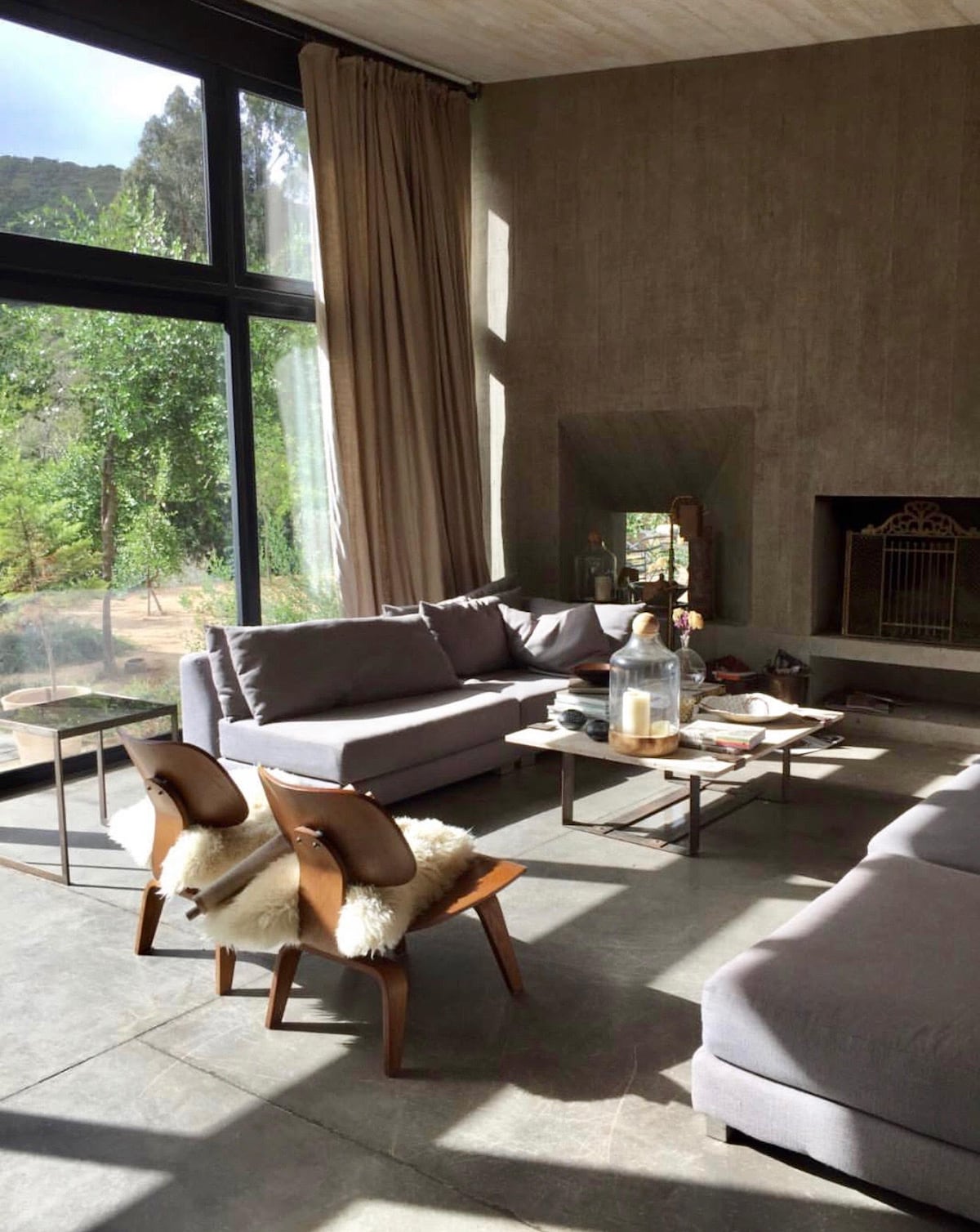 A modern living area is illuminated by natural light streaming through large glass windows. Two comfortable sofas are positioned around a low coffee table, which holds decorative items. Wooden armchairs with soft coverings provide extra seating, while a fireplace adds to the cozy ambiance.