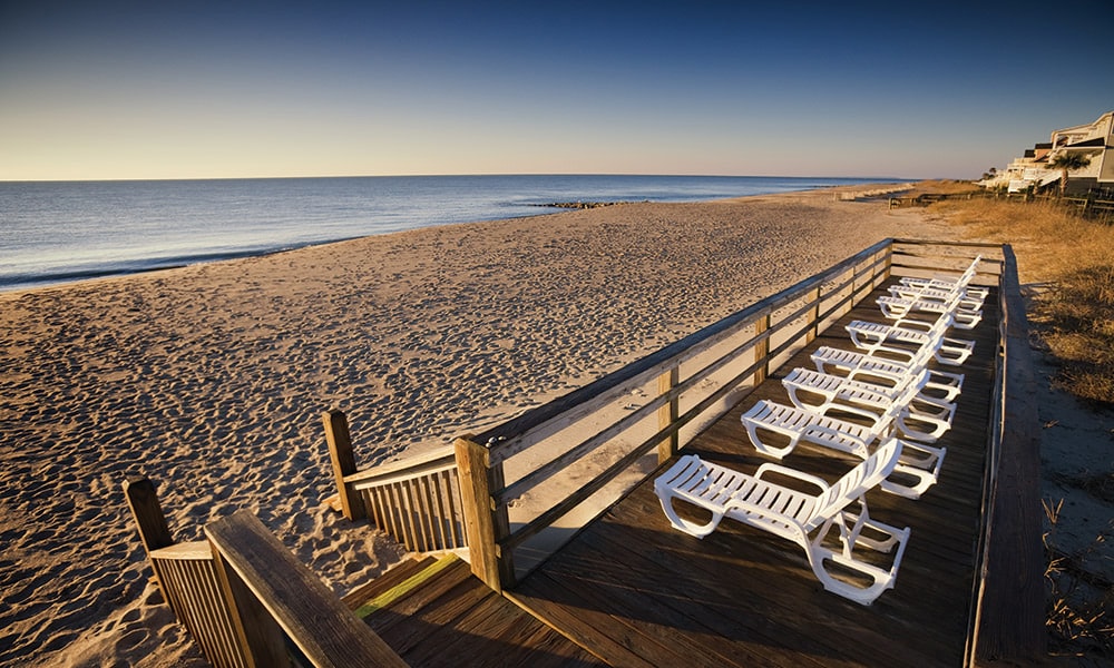 A wooden boardwalk extends toward the beach, lined with multiple white lounge chairs positioned for sunbathing. The calm ocean is visible in the distance under a clear sky, while smooth sand stretches along the shoreline.