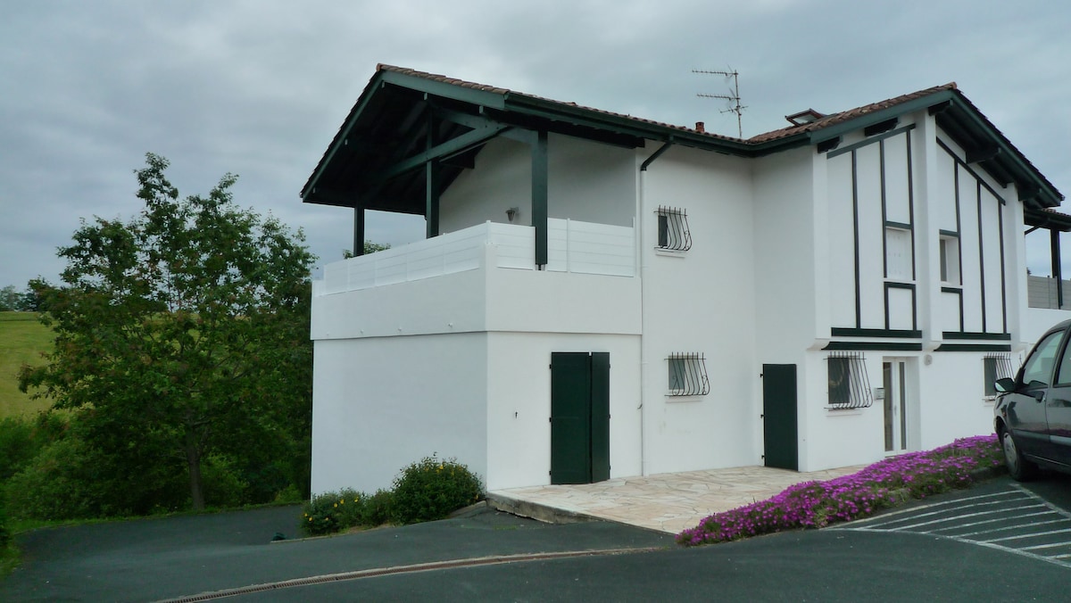 A two-story building is presented, featuring a traditional exterior with white and dark accents. A spacious balcony is visible on the upper level, bordered by decorative railings. Surrounding greenery and flowering plants enhance the tranquil setting, while a parked car is visible nearby.