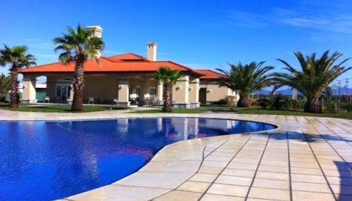 A large outdoor pool is framed by a spacious tiled deck and palm trees. The house features a sloped red tile roof and numerous windows. Clear blue skies provide a bright backdrop, enhancing the open and inviting landscape.