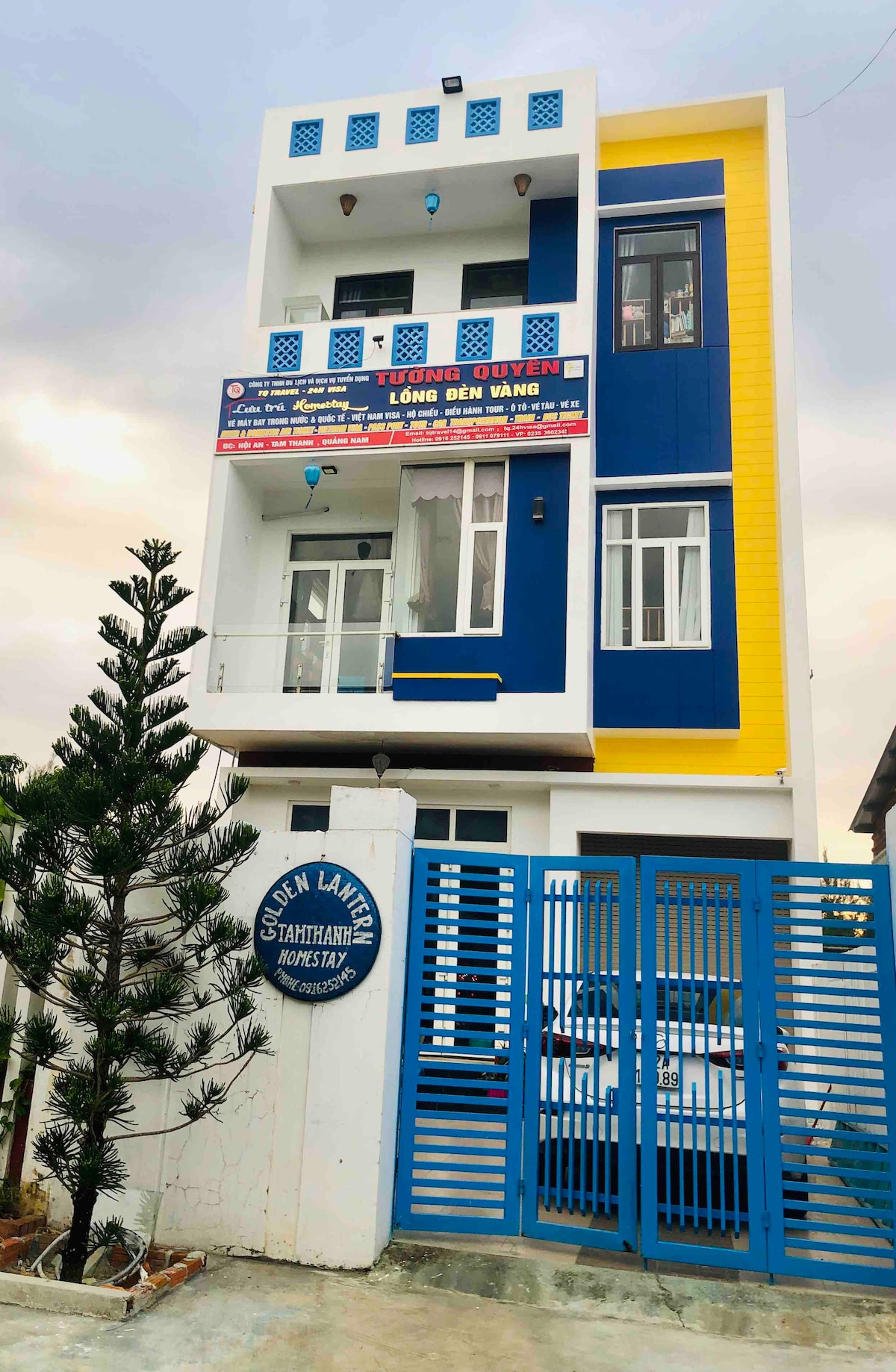 The homestay showcases a three-story building painted in a vibrant blue and yellow color scheme. A tree is positioned nearby, while a secure blue gate provides access. The design reflects a modern style, harmonizing with its surroundings.
