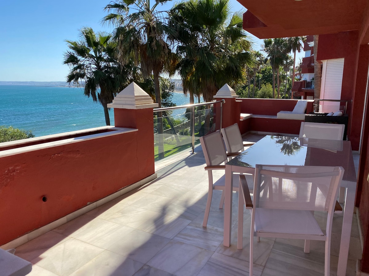 A spacious terrace is visible, featuring a large glass table surrounded by several white chairs. Palm trees frame the view of the beach and sea beyond, with the sunlight reflecting off the water. The exterior walls are painted in a warm red tone.