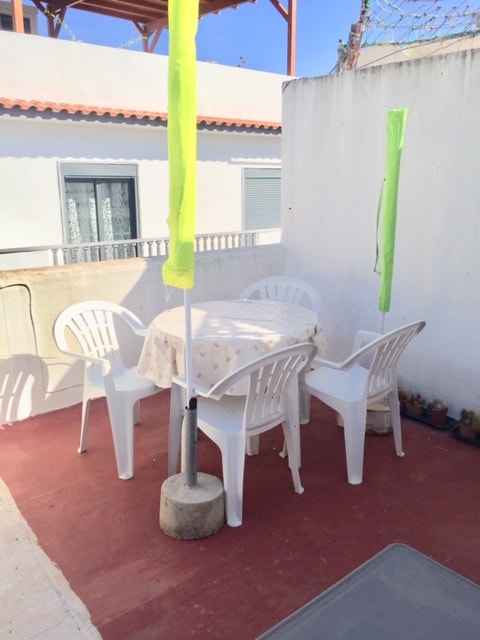 A small terrace is shown with a round table and four white plastic chairs. Bright green flags carefully anchored are placed in the corners. The space is enclosed by low walls, and the sunny sky is visible above, creating a comfortable outdoor setting.