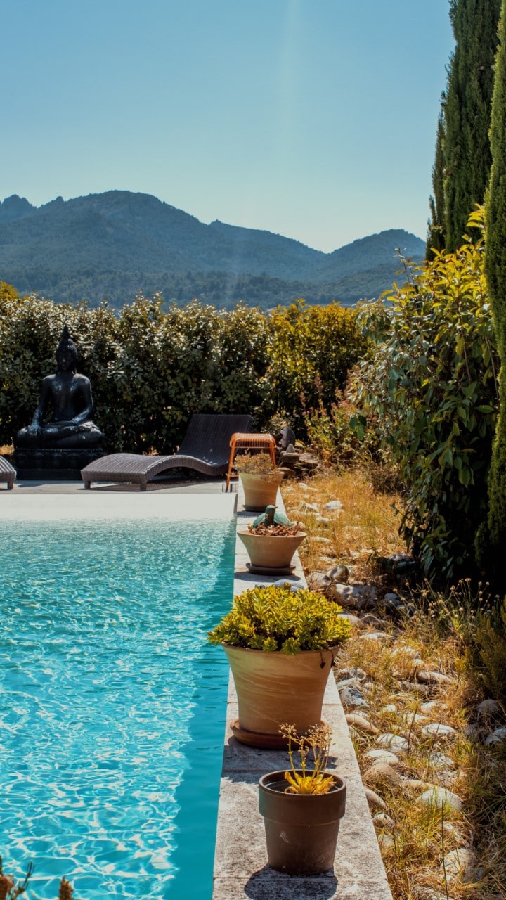 A serene outdoor pool area is framed by potted plants and decorative statues. Comfortable lounge chairs are positioned beside the water. Rolling hills are visible in the background, creating a tranquil atmosphere. Bright sunlight enhances the clear blue of the pool.