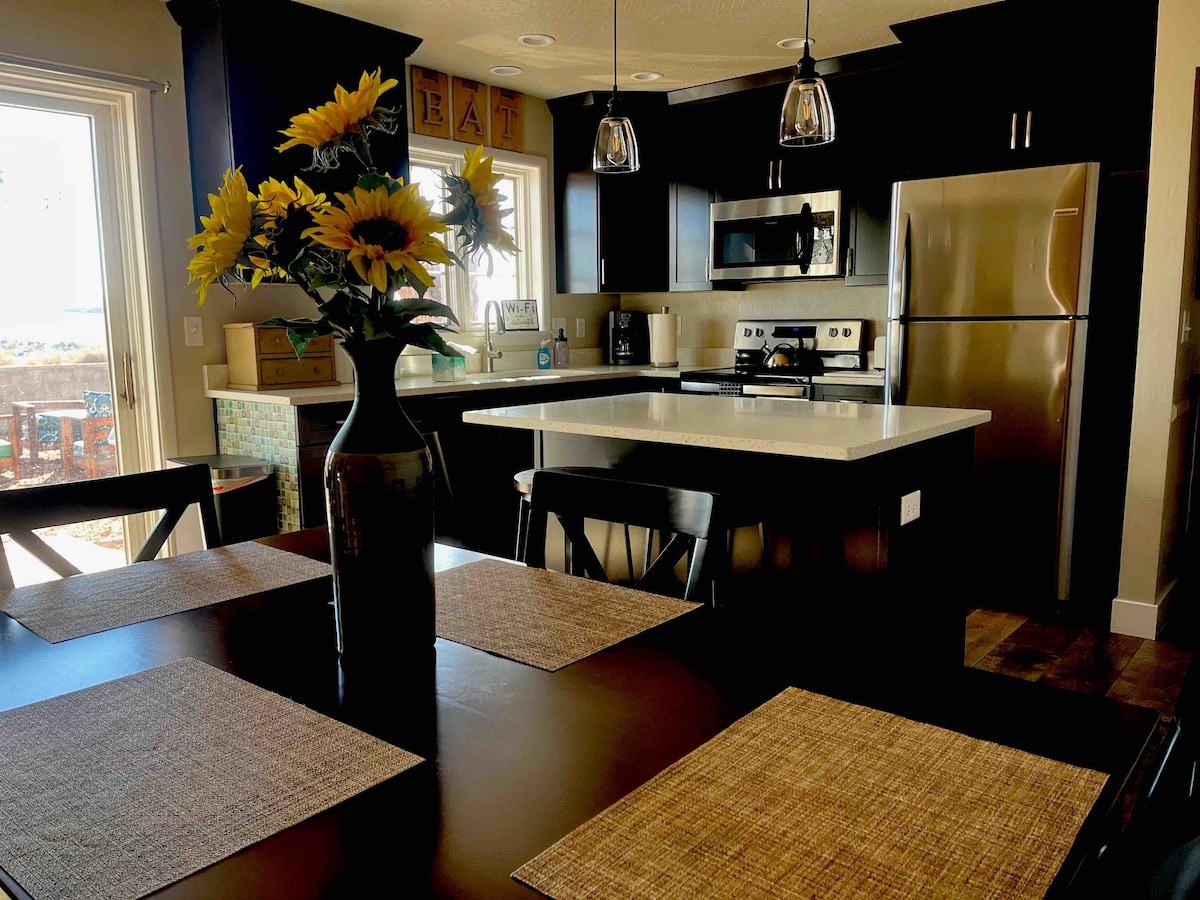 A modern kitchen is displayed with dark cabinetry and a spacious island countertop. Sunflowers are arranged in a tall vase on the dining table, complemented by woven placemats. The kitchen includes stainless steel appliances and an inviting view through a nearby window.