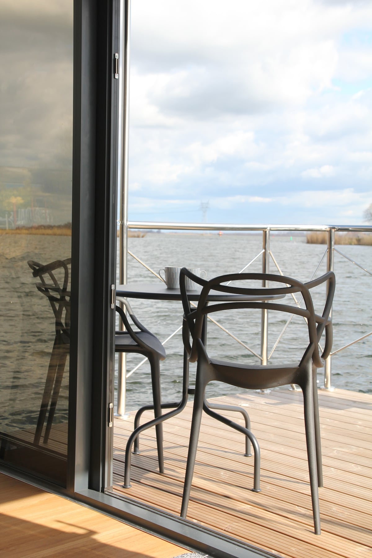 A spacious balcony overlooks the water, featuring a small table with two chairs. Glass doors provide a seamless transition from the interior, reflecting the view of the serene water and cloudy sky.