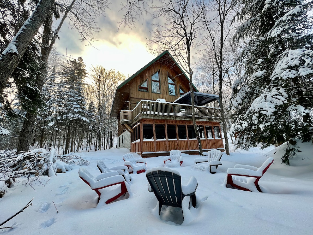 A charming chalet is nestled among snow-covered trees, featuring a spacious deck and dark wooden exterior. Circular seating made of various colored chairs is arranged in the snowy ground, providing a cozy gathering space. The soft light of dawn filters through the trees.