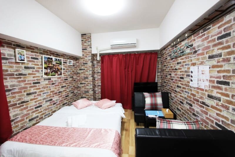 The room features a double bed with pink pillows and crisp white linens, accompanied by a cozy sofa. The brick-patterned wallpaper adds texture, while red curtains frame the windows, creating a comfortable ambiance. A small TV is positioned against the wall, enhancing the relaxed setting.