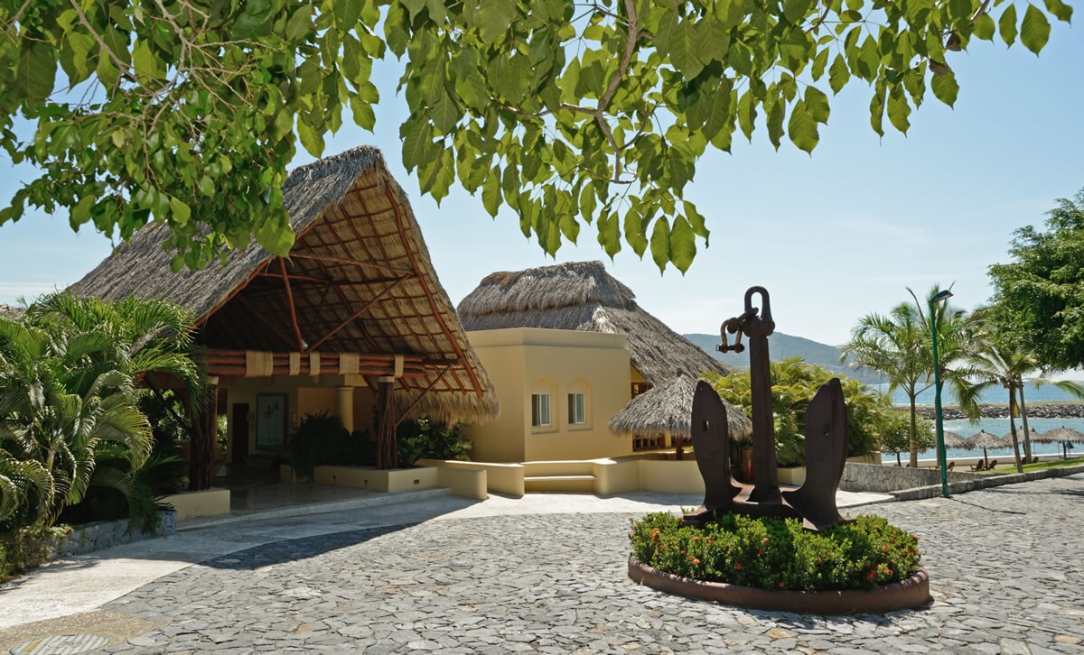 A charming entrance features a thatched roof structure surrounded by lush greenery, with stone pathways leading to the villa. A large anchor sculpture is placed prominently in the foreground, enhancing the coastal ambiance of the location.