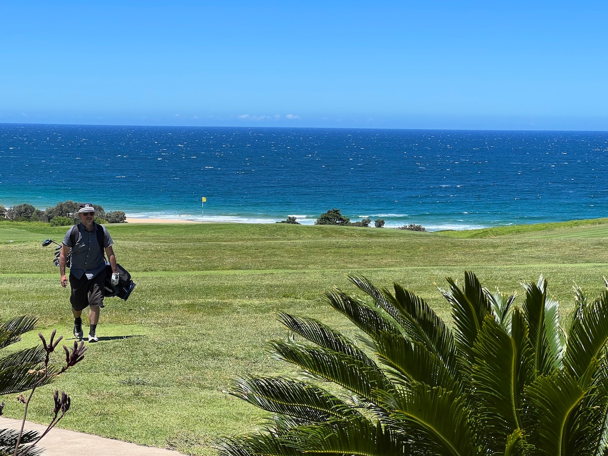 A golfer is seen walking across a manicured green, carrying golf clubs. In the background, a serene blue ocean meets the horizon under a clear sky, while lush greenery frames the scene, enhancing the natural surroundings.