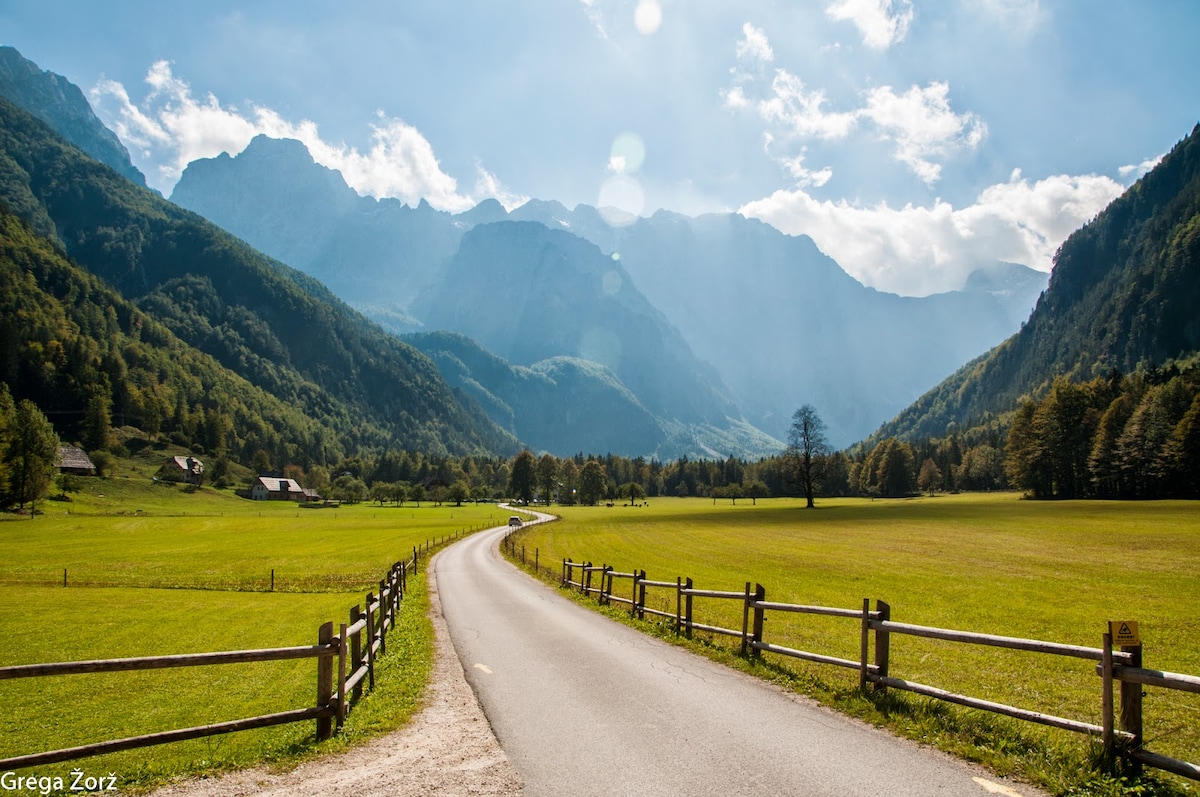 A winding road leads through a lush green meadow, flanked by a wooden fence. Majestic mountains rise in the background, partially obscured by soft clouds, with sunlight filtering through, creating a serene atmosphere in the Upper Savinjska Valley.