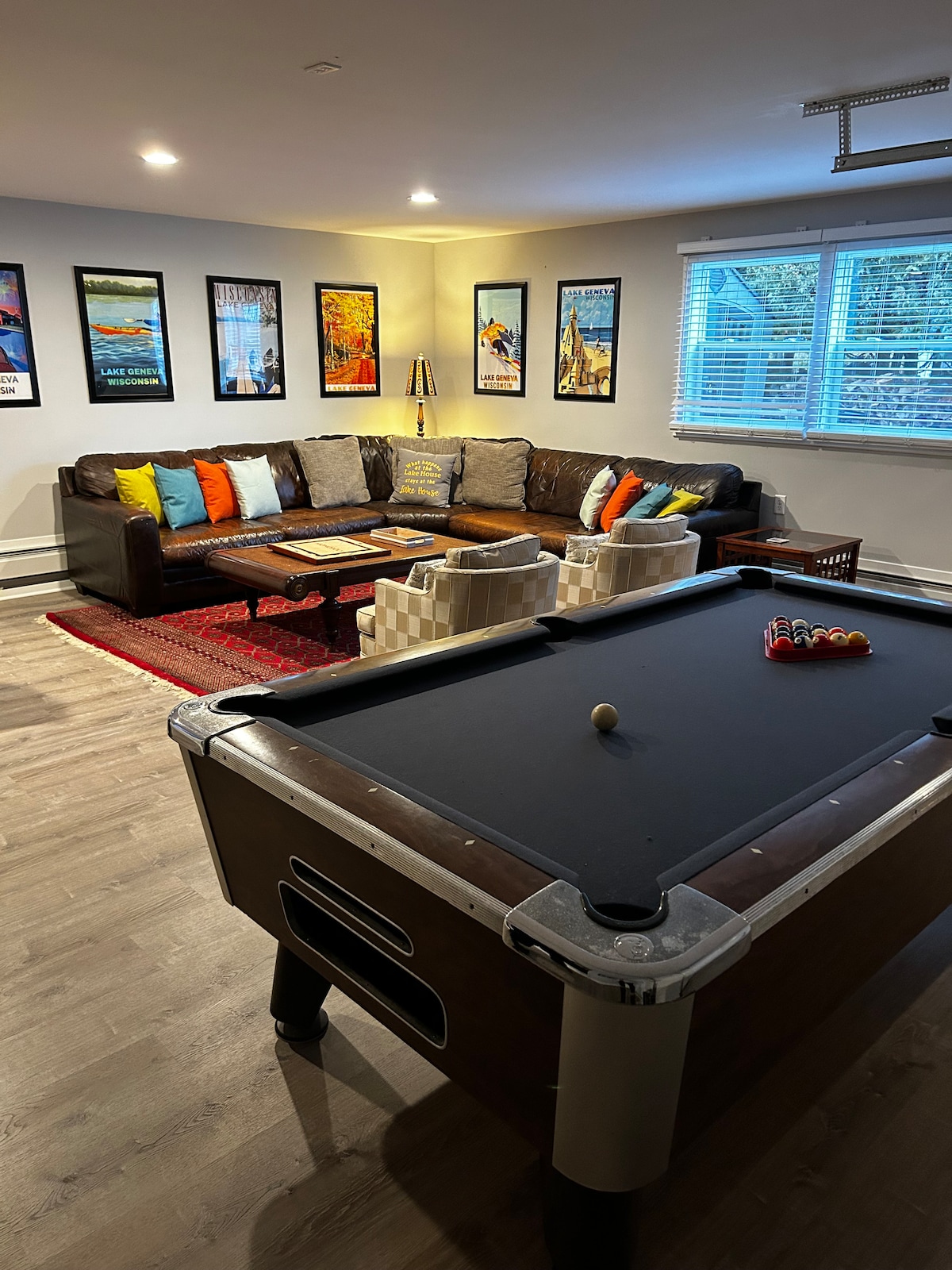 A modern game room is presented, featuring a pool table at the forefront. A spacious, comfortable couch adorned with colorful pillows lines the wall, surrounded by framed artwork. Natural light enters through a window, illuminating the room's inviting atmosphere.