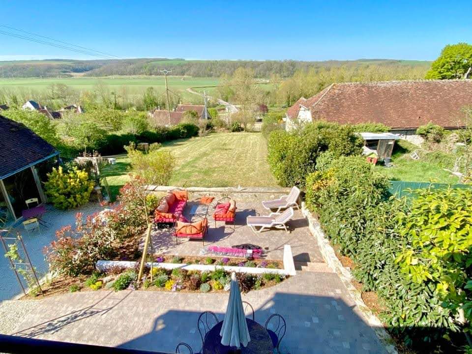 A spacious outdoor area is depicted, featuring comfortable seating arranged around a vibrant garden. Lush greenery surrounds the space, with views of rolling hills in the background. A terrace is visible, offering a relaxing atmosphere for guests to enjoy.