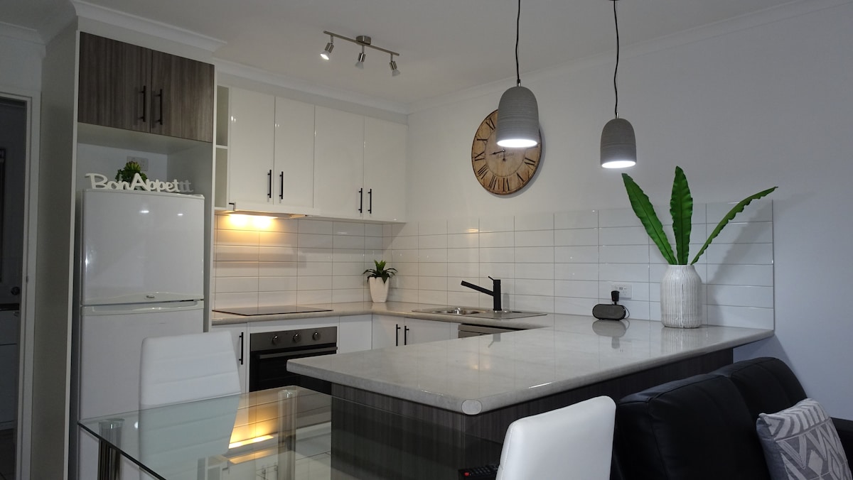 A modern kitchen is showcased, featuring sleek cabinetry and a spacious stone countertop. Pendant lights hang above, illuminating the area. A plant in a white vase adds a touch of greenery, while a wall clock provides a classic accent. The dining table is visible, complementing the kitchen space.