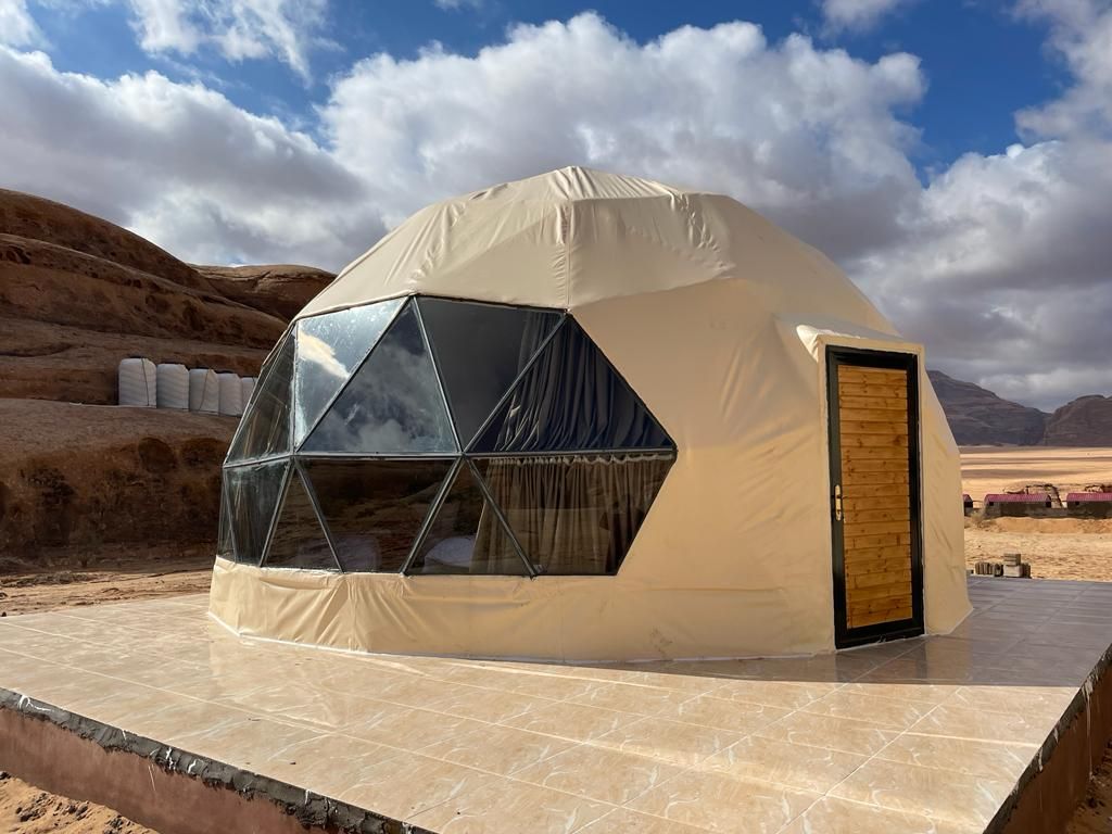 A geodesic dome structure is positioned on a stone patio, featuring large triangular glass panels that offer views of the surrounding desert landscape. A wooden door with a natural finish provides entrance. The dome's exterior is covered in a beige fabric, complementing the earthy tones of the environment.