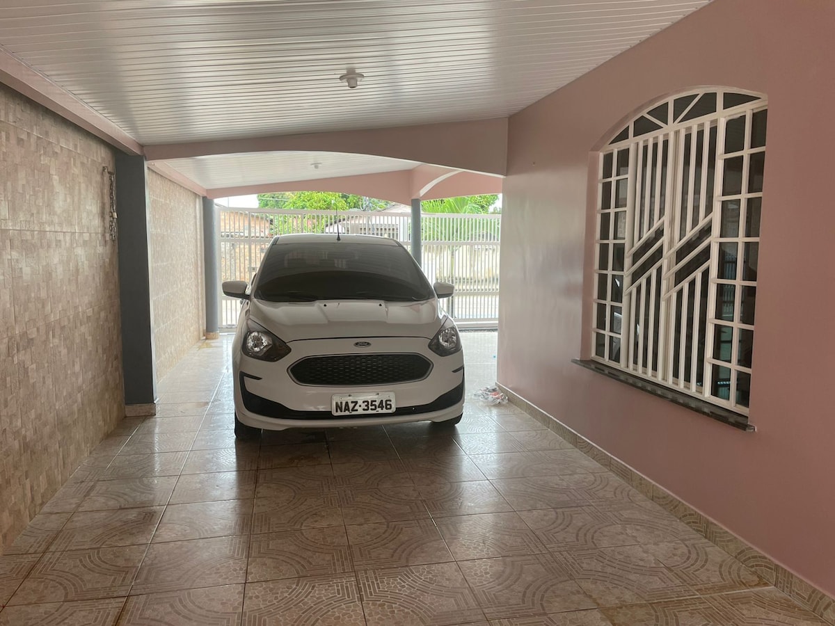 A covered parking area is visible featuring a white vehicle positioned in front. The space is framed by beige tiled walls and a decorative window with white bars allowing natural light. The entrance's safety features include an electric gate and a secure environment.
