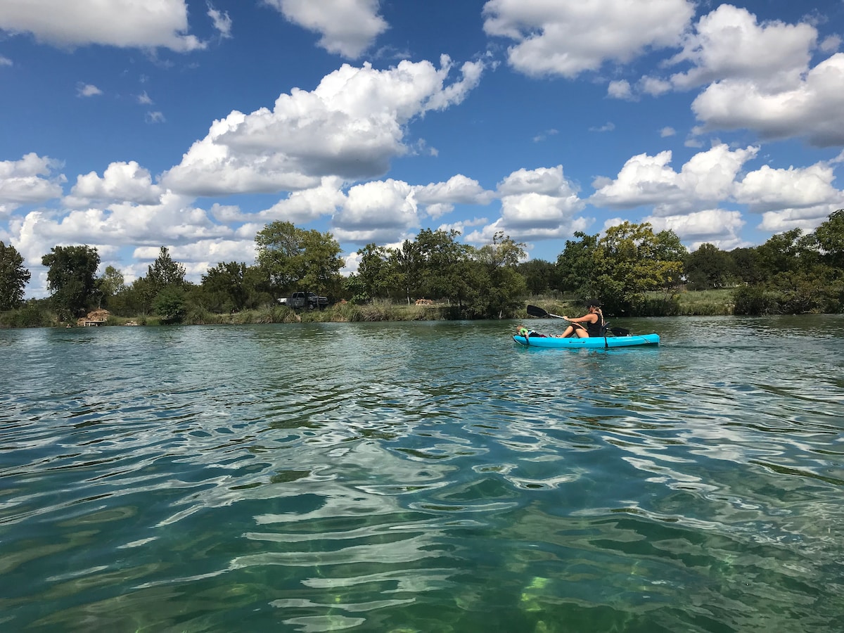 A kayak is seen gliding across the clear water of the Nueces River, with a person paddling. Lush greenery lines the riverbanks under a bright blue sky dotted with fluffy white clouds.